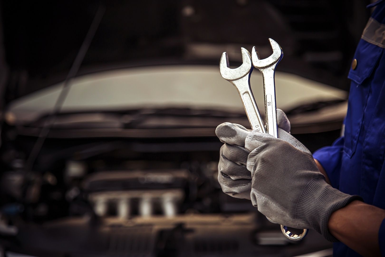 Eine Person in blauer Arbeitskleidung und Handschuhen hält zwei Schraubenschlüssel über dem geöffneten Motorraum eines Autos
